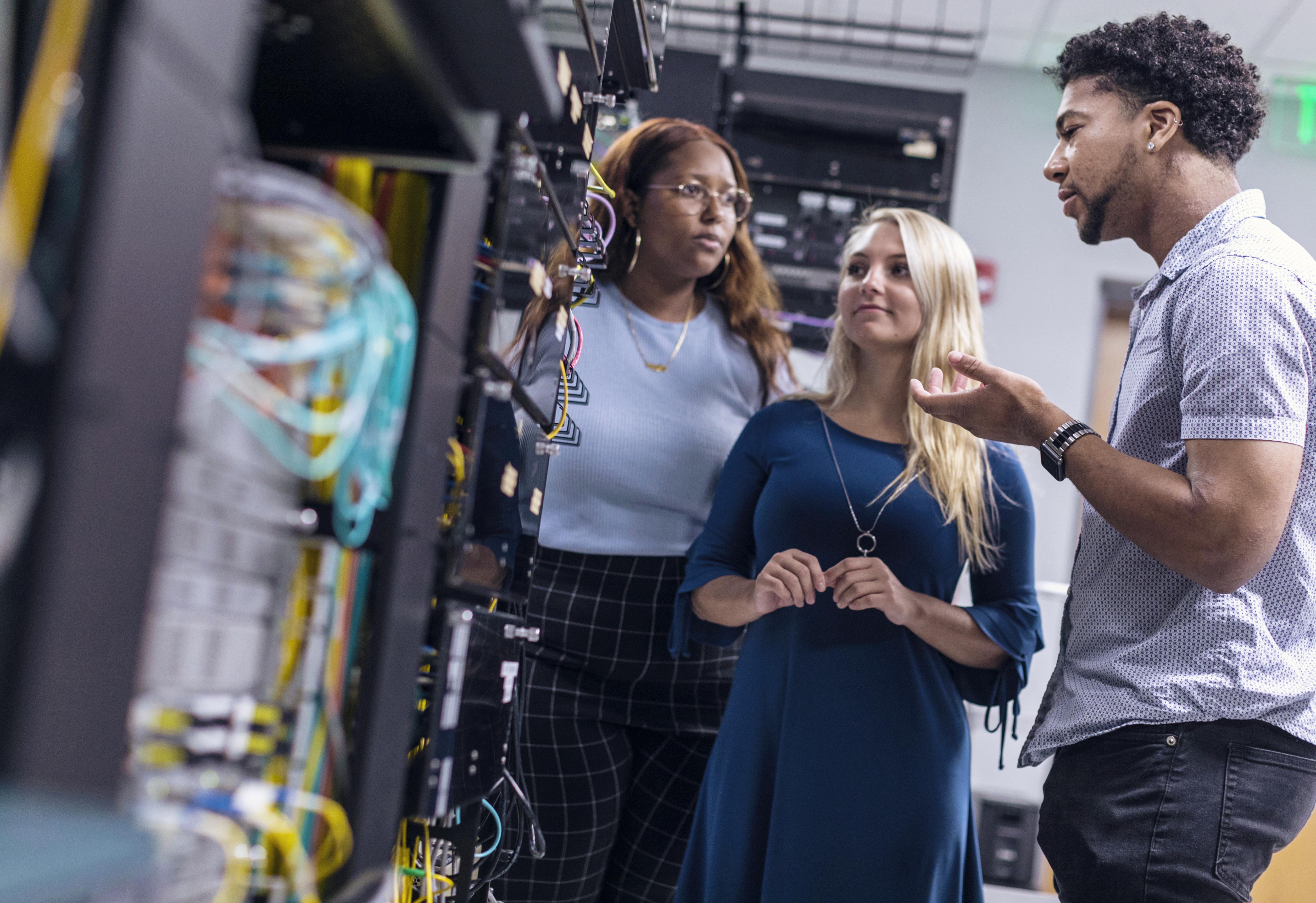 A group of three students are standing around electrical equipment in the cyber security lab